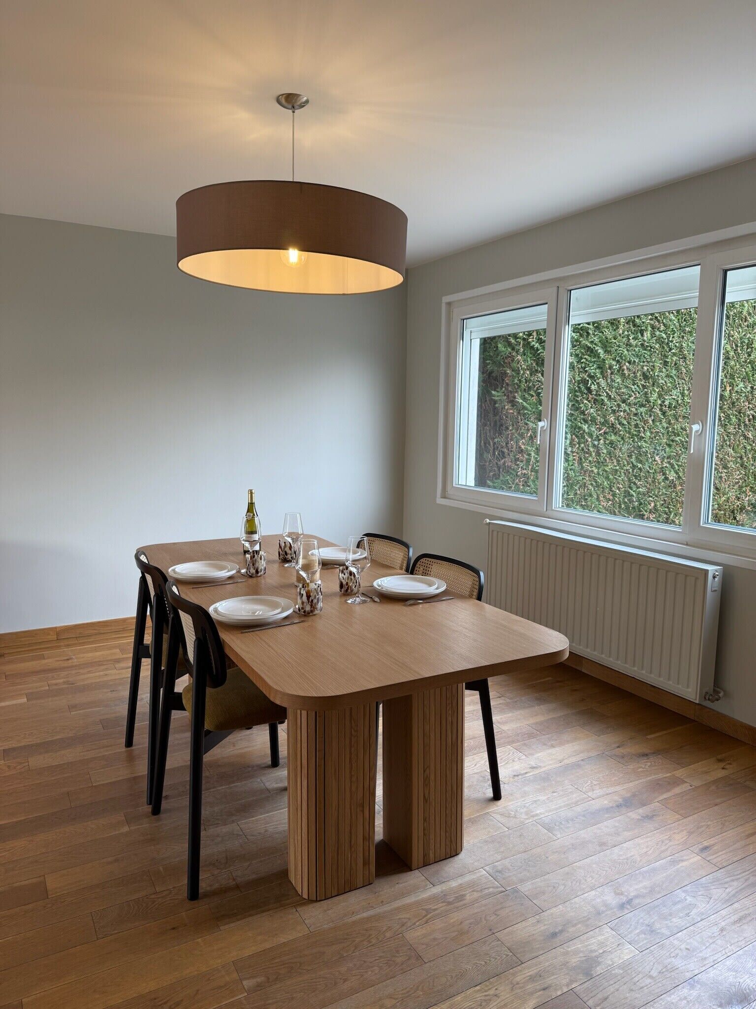 Ein Essbereich mit einem Holztisch, gedeckt für vier Personen, einer Hängelampe und einem Fenster mit Blick ins Grüne.