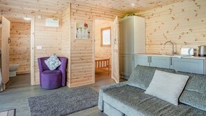 Interior acogedor de una cabaña con sofá, una silla morada, una mesita y una pequeña cocina.