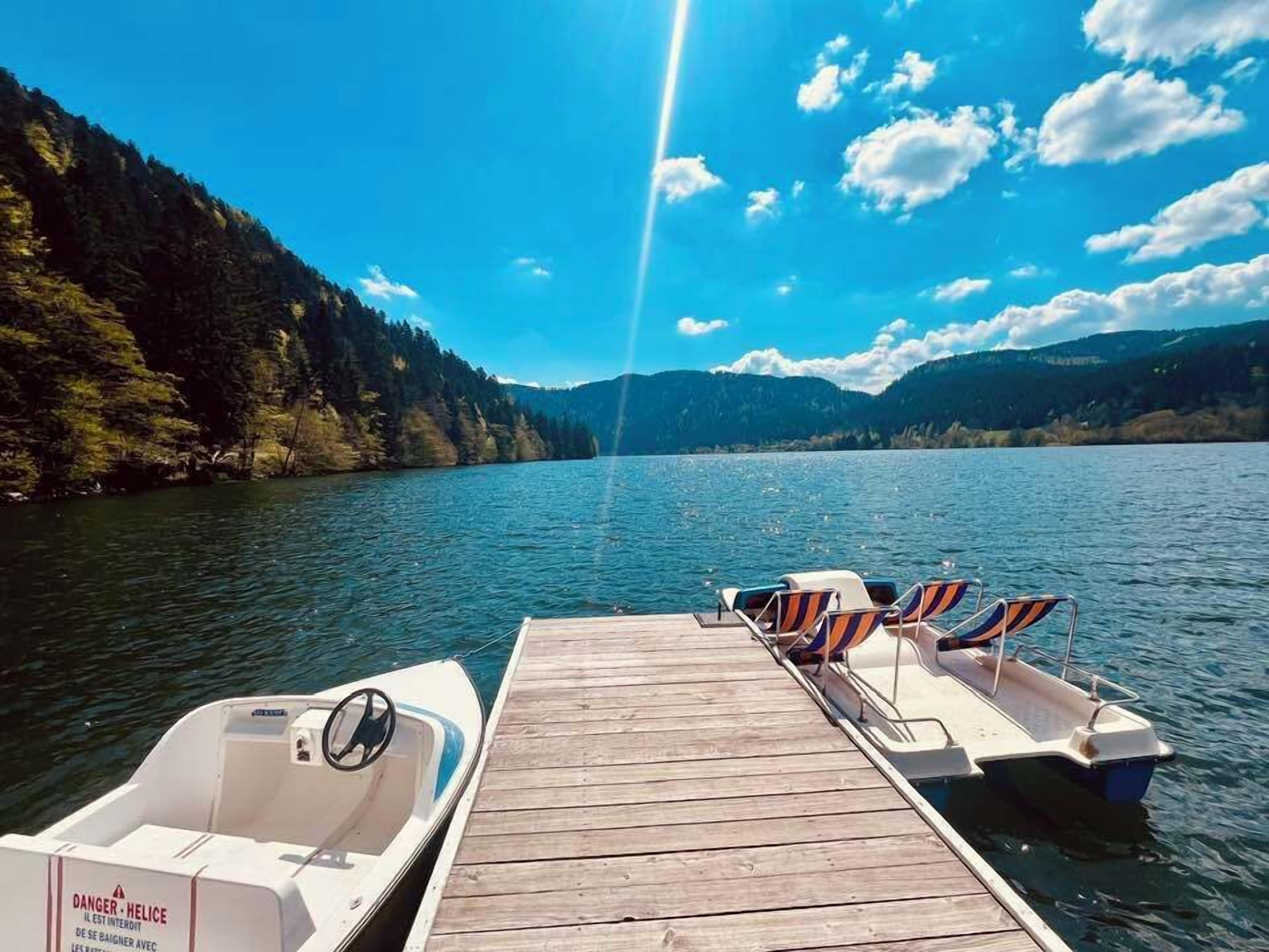 Een houten steiger met een roeiboot en lounge stoelen aan een rustig meer, omgeven door beboste heuvels.
