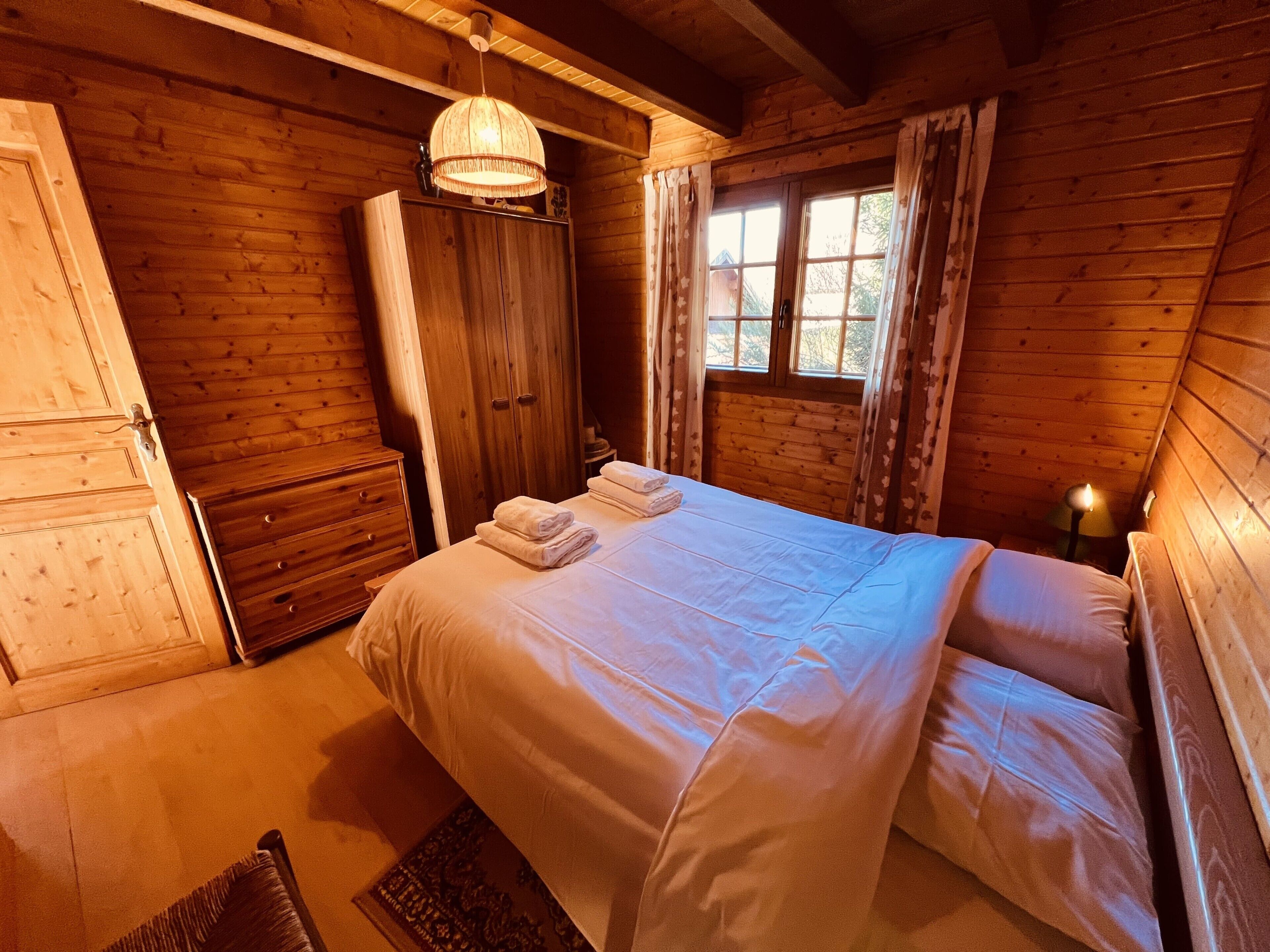 A wooden cabin bedroom with a bed, a wardrobe, and a window with curtains.