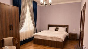 A bedroom with a wooden bed, a nightstand, a wardrobe, a chair, a table with water bottles, and a potted plant. - Silk Nest Hotel (Bukhara)