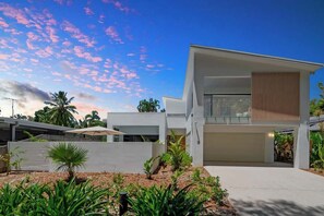 A modern two-story house with a flat roof, a garage, and a landscaped garden with tropical plants.