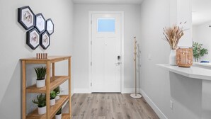 A hallway with a white door, a wooden console table with plants, a mirror, and a wall-mounted hexagonal display.
