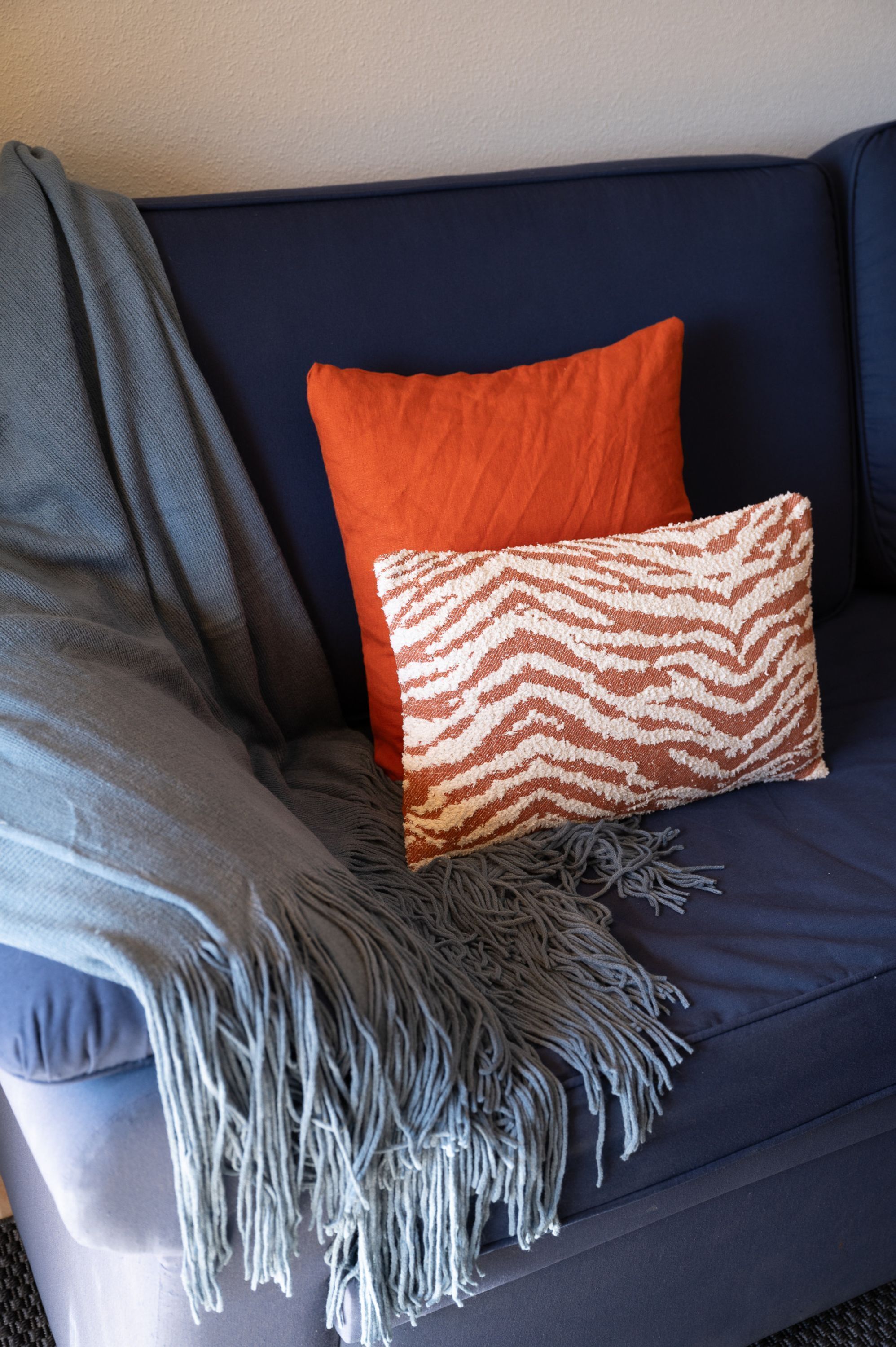 A dark blue sofa with a grey throw blanket and two pillows, one orange and one with a chevron pattern.