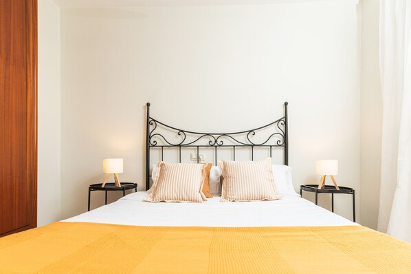 A neatly made bed with a white duvet and two striped pillows, flanked by two bedside tables with lamps, against a plain wall.