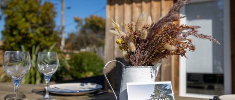 A wooden table with a metal pitcher holding dried plants, two empty wine glasses, and a postcard with the text "CocoNR".
