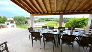 An outdoor patio with a wooden ceiling, a dining table set for six, and a view of a swimming pool and greenery. - Maison Roquebeauresse - Piscine privée (Pougnadoresse)