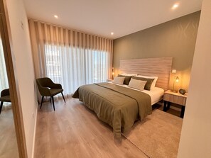 Un dormitorio moderno con una cama grande, una silla, una mesita de noche con lámpara y una ventana con cortinas.