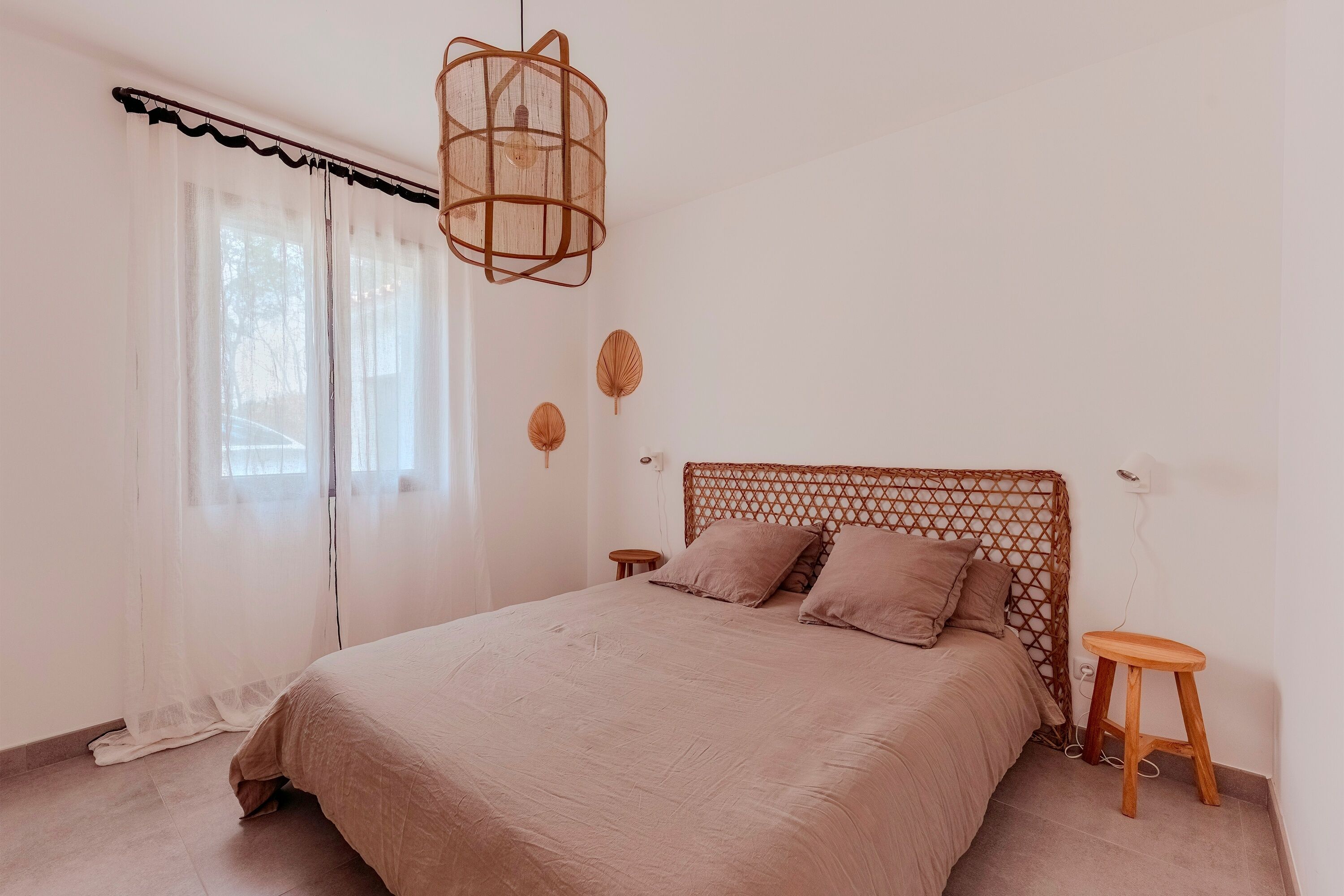 Ein Schlafzimmer mit einem Bett, einem Holzkopfende, einer Hängeleuchte, einem kleinen Hocker und einem Fenster mit transparenten Vorhängen.