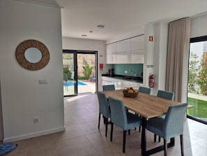 Une cuisine moderne avec des meubles blancs, un coin repas avec une table en bois et des chaises bleues, et une vue sur une piscine à l’extérieur.
