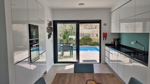 Une cuisine moderne avec des meubles blancs, une table à manger avec des chaises, une vue sur une piscine extérieure et un mur en pierre.