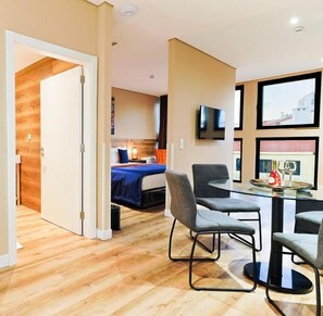 A modern hotel room with a wooden floor, a bed with a blue cover, a glass table with chairs, and a flat-screen TV mounted on the wall. - CONDE13 Aparthotel Boutique  (Lisboa)