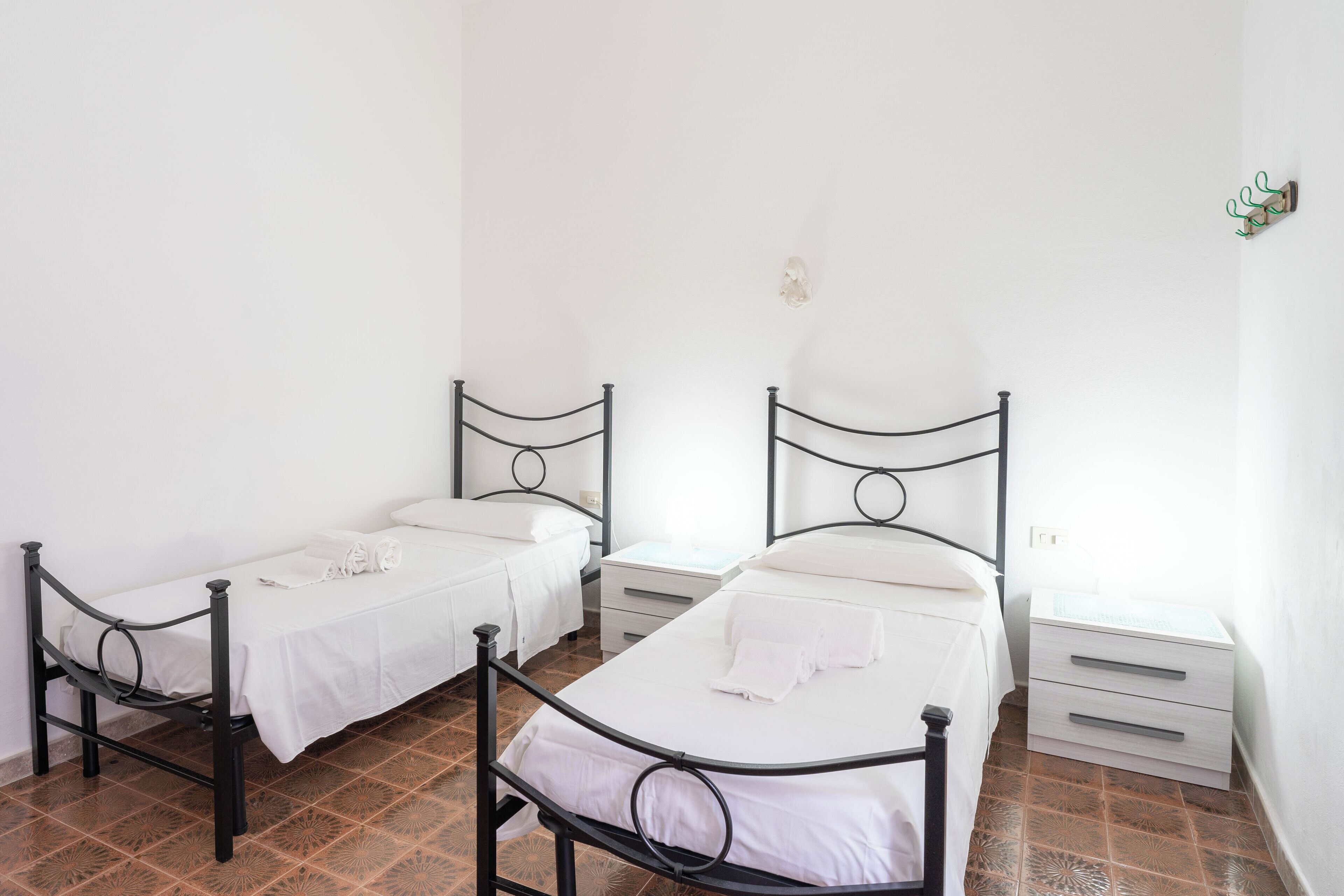 Deux lits simples avec des structures en métal et du linge de lit blanc, une petite table de chevet avec des tiroirs et un luminaire fixé au mur.
