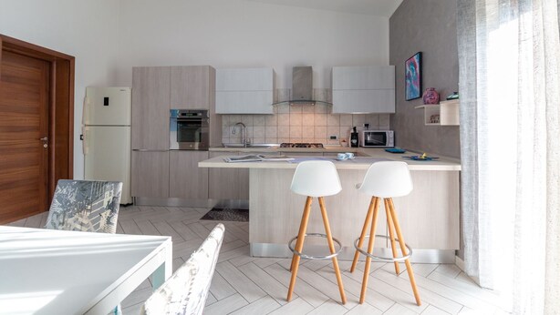 Eine moderne Küche mit Holzausstattung, einem eingebauten Backofen, einer Mikrowelle und einem Gasherd. Zwei weiße Barhocker mit Holzbeinen ergänzen das Bild.