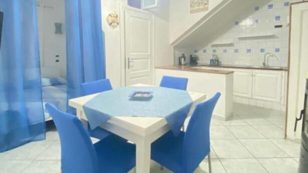 Un comedor con una mesa blanca y sillas azules, una cocina con armarios blancos, y una escalera que sube a otro nivel.