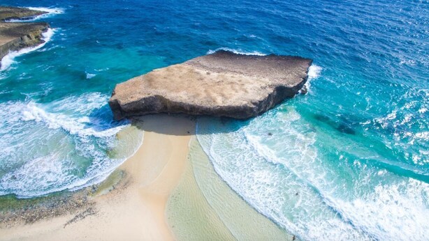 Eine Vogelperspektive zeigt einen Strand mit einer großen Felsformation in der Mitte, umgeben von türkisblauem Wasser und Sandufern.