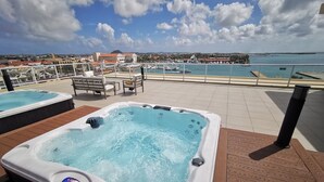 Ein Whirlpool auf einer Holzterrasse mit Blick auf das Meer und Gebäude im Hintergrund.