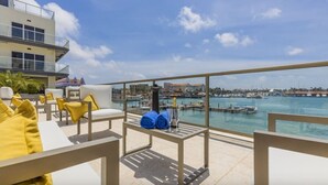 Eine Dachterrasse mit weißen Liegestühlen und gelben Kissen mit Blick auf eine Marina, in der Boote und Gebäude liegen.