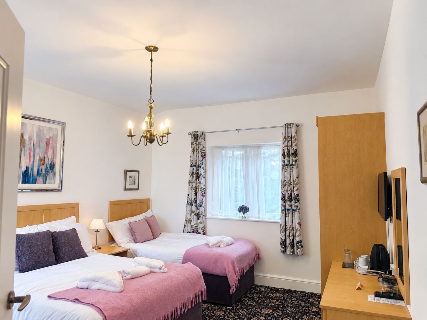 Habitación de hotel con dos camas, un candelabro, un ventanal con cortinas florales, un escritorio de madera y un televisor.