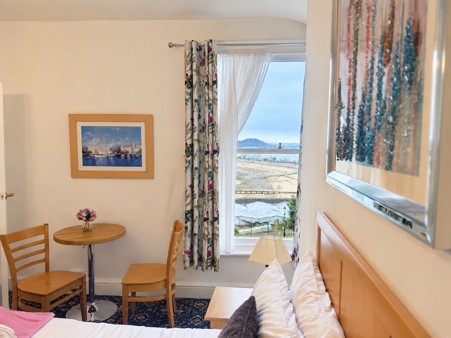 Habitación de hotel con cama, escritorio, dos sillas, una pequeña mesa con un jarrón de flores, un cuadro en la pared y una ventana con vistas al paisaje.