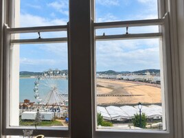 Vista a través de una ventana mostrando una zona costera con playa, una noria grande y una hilera de edificios.