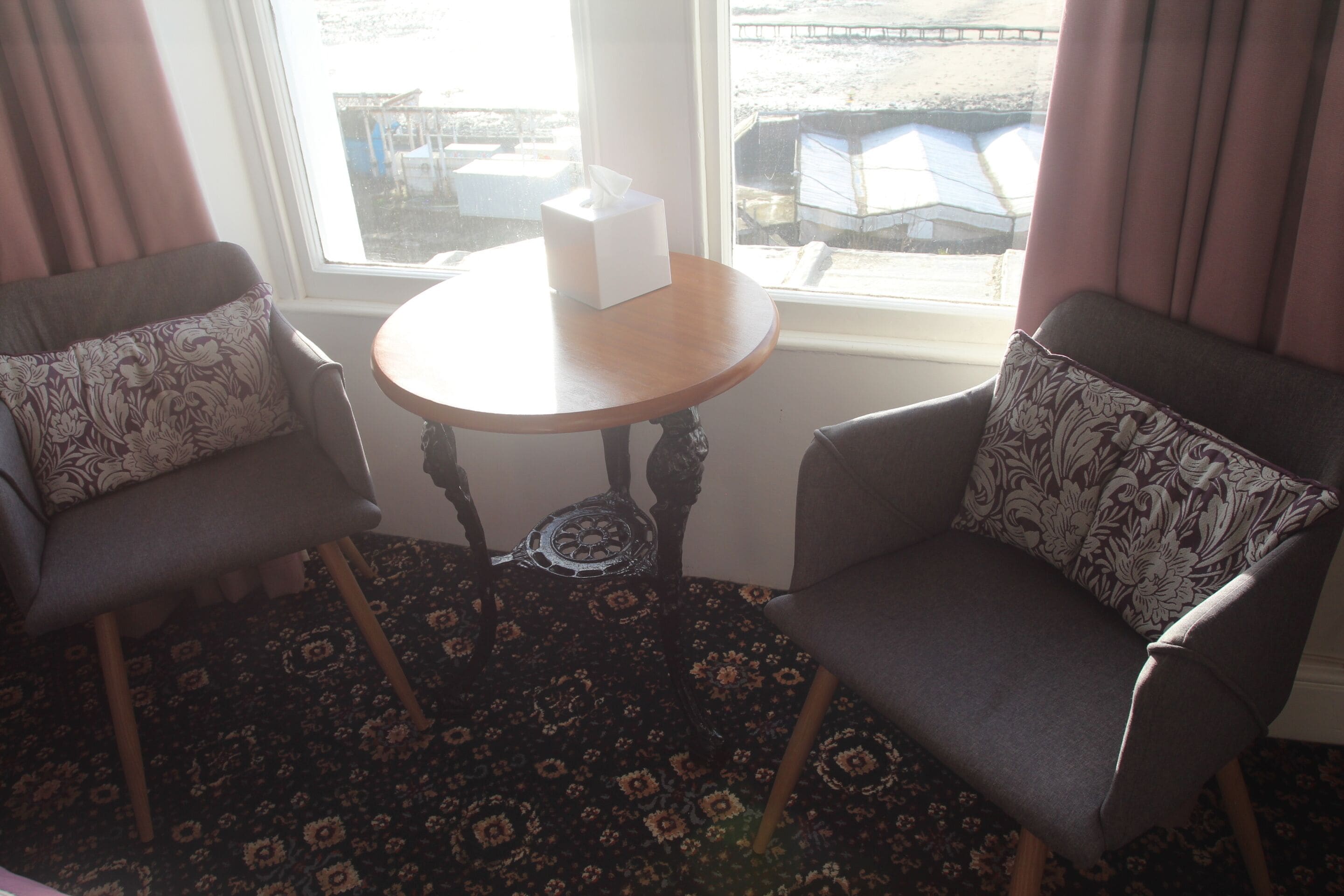 A room with two armchairs, a small round table with a tissue box, and a view of a dock and boats through a window.