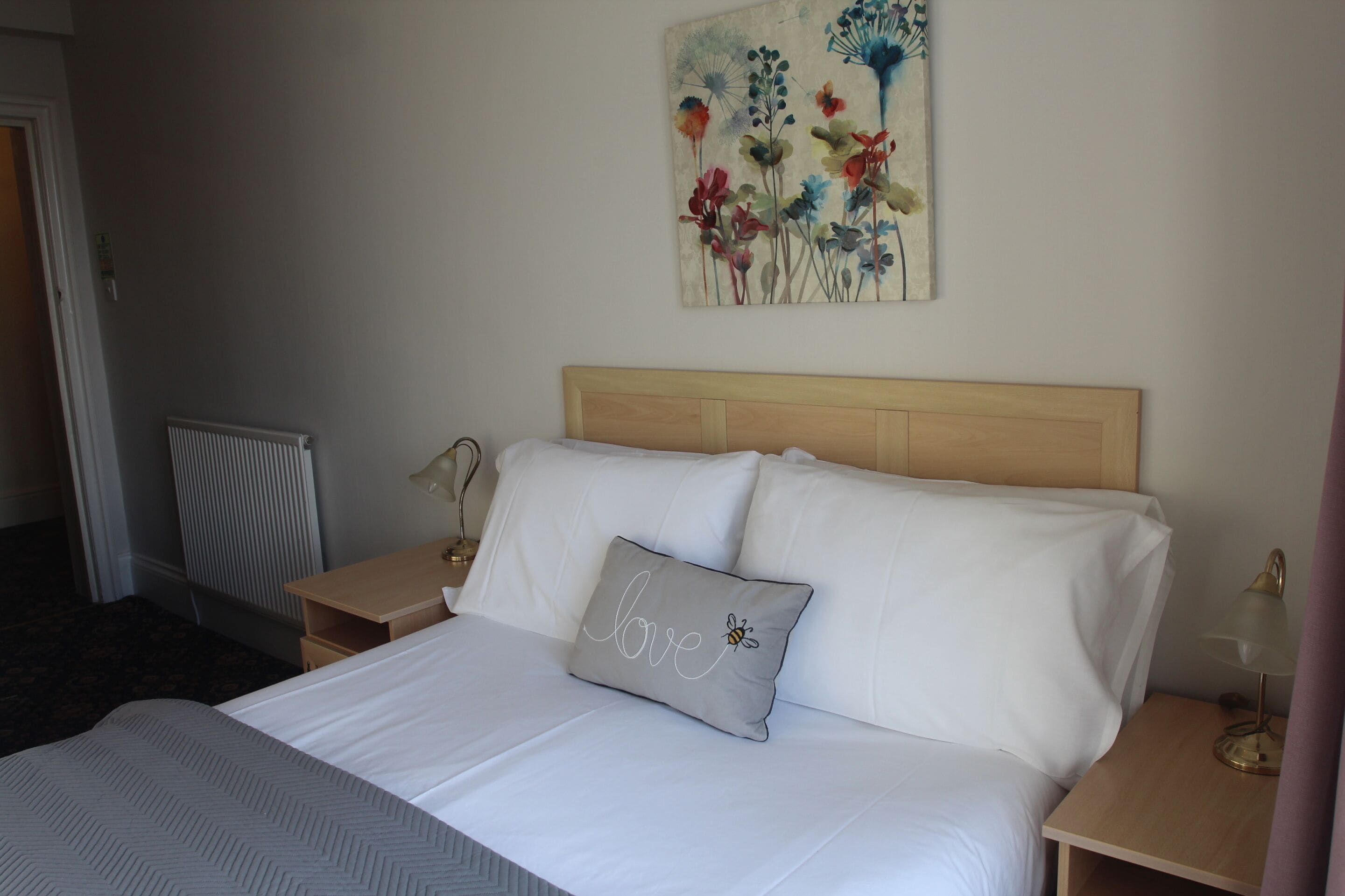 Una cama bien hecha con sábanas blancas y una almohada con la palabra "love" y un diseño de abeja. Se observa un cabecero de madera, una mesita de noche con una lámpara y un cuadro de flores en la pared.