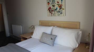Una cama bien hecha con sábanas blancas y una almohada con la palabra "love" y un diseño de abeja. Se observa un cabecero de madera, una mesita de noche con una lámpara y un cuadro de flores en la pared.