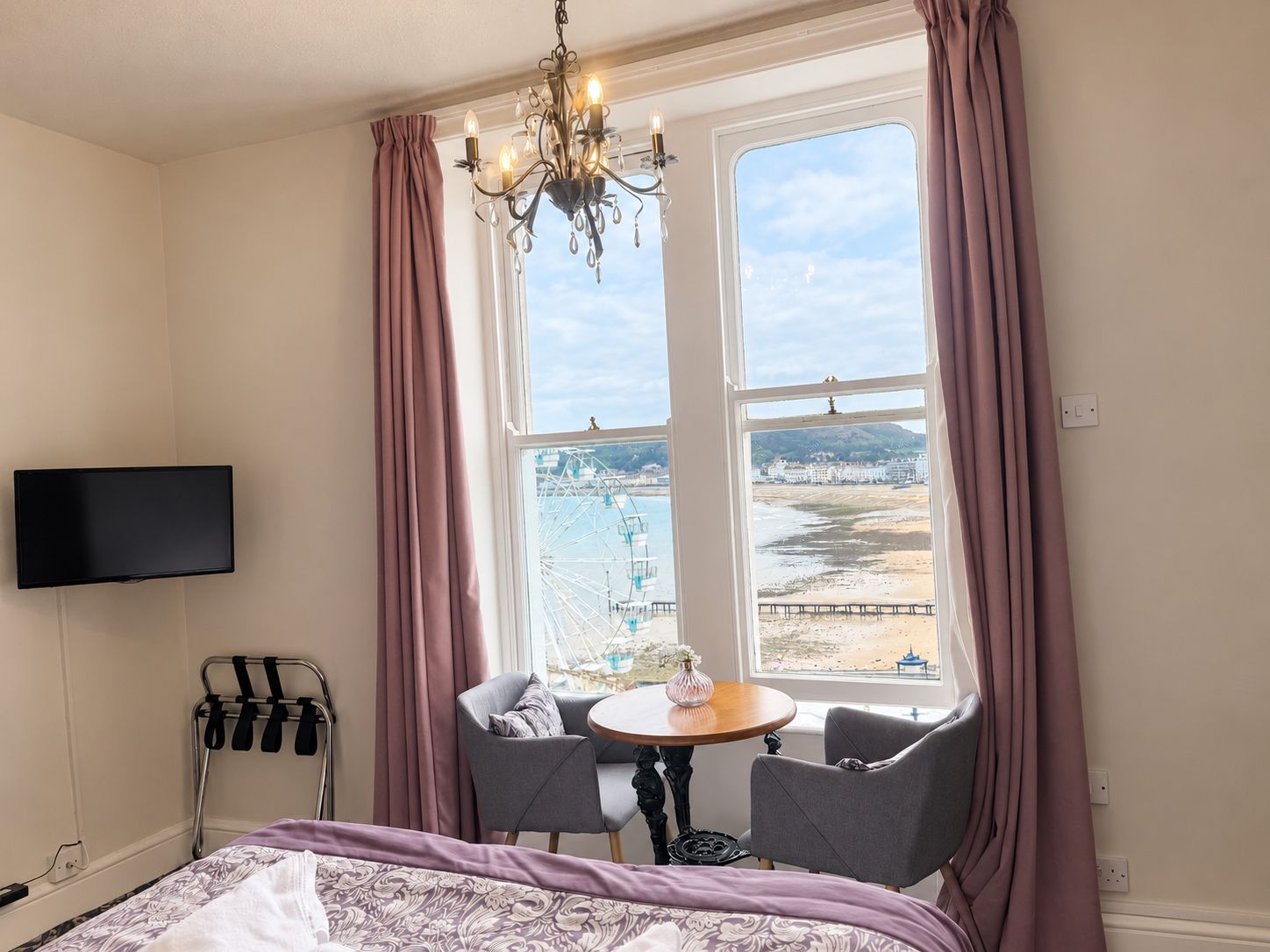 Habitación de hotel con cama, televisión montada en la pared, mesa con dos sillas y un ventanal con cortinas.
