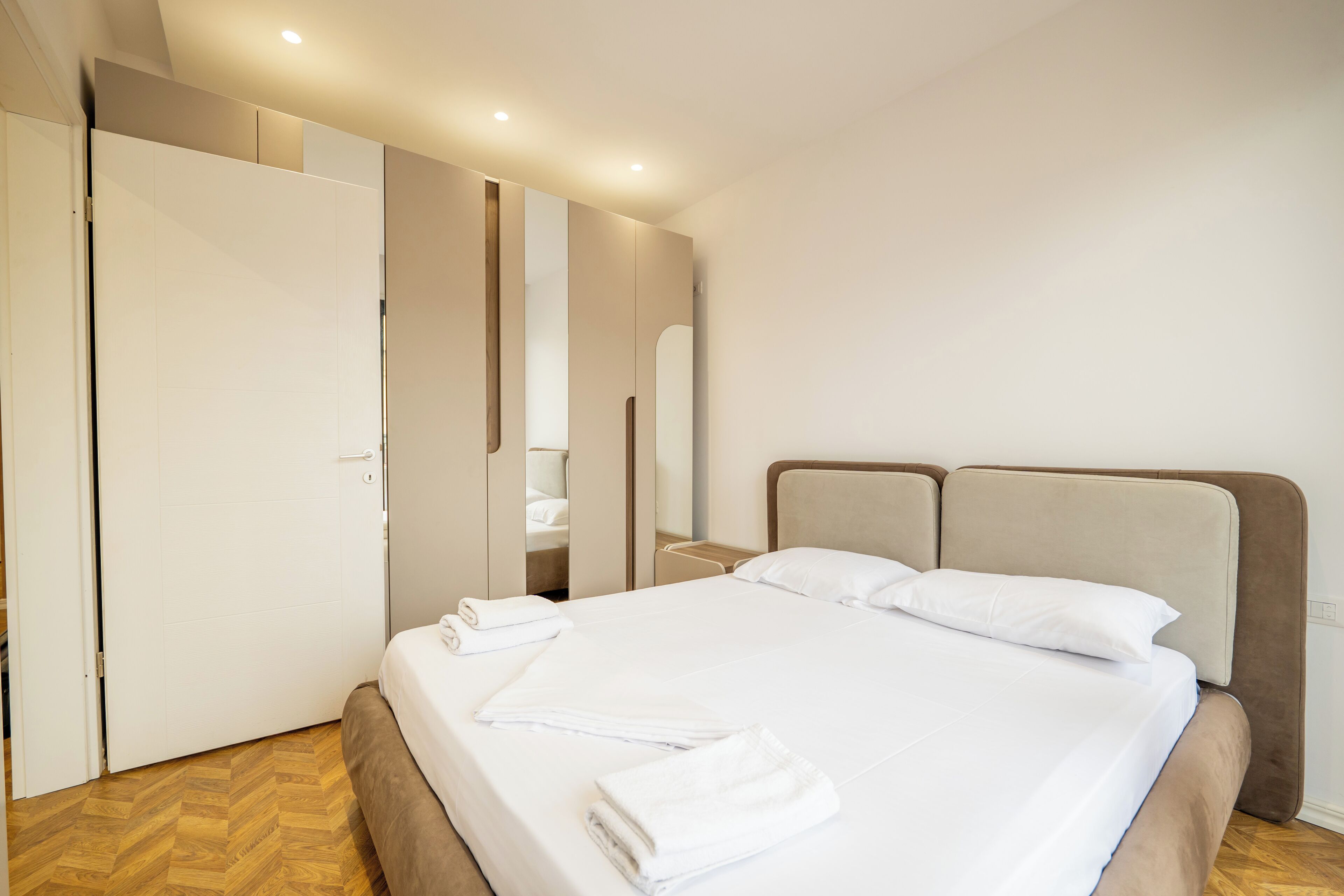 Una cama bien hecha con almohadas y toallas dobladas, suelo de madera y una puerta cerrada.