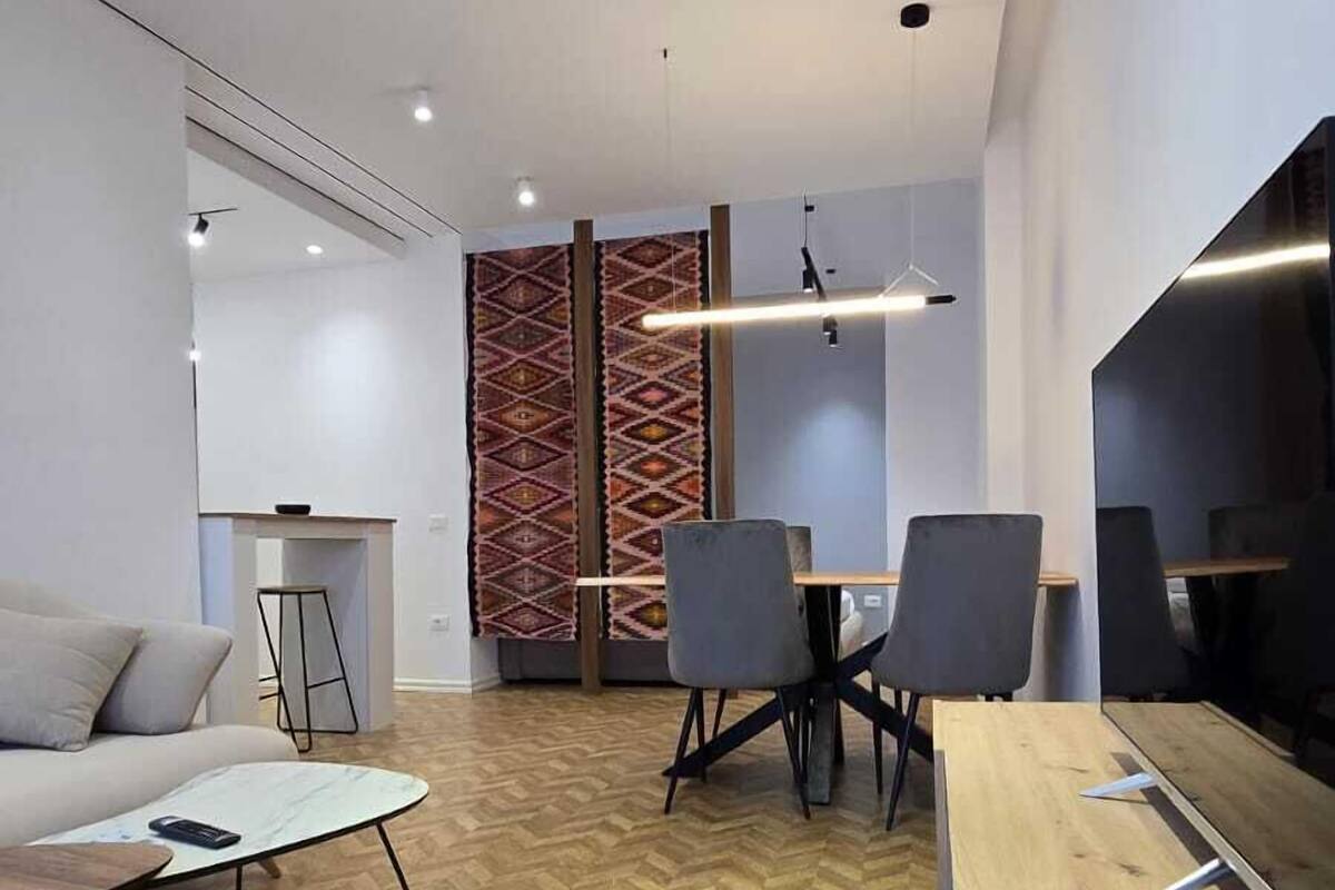 A modern, well-lit room with a herringbone wood floor, a sofa, a dining table with chairs, and a kitchen area with a stove.