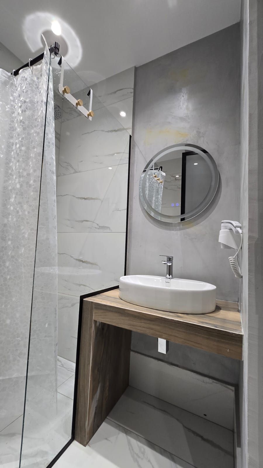 A modern bathroom with a glass shower enclosure, a round mirror, and a wooden vanity with a white sink.