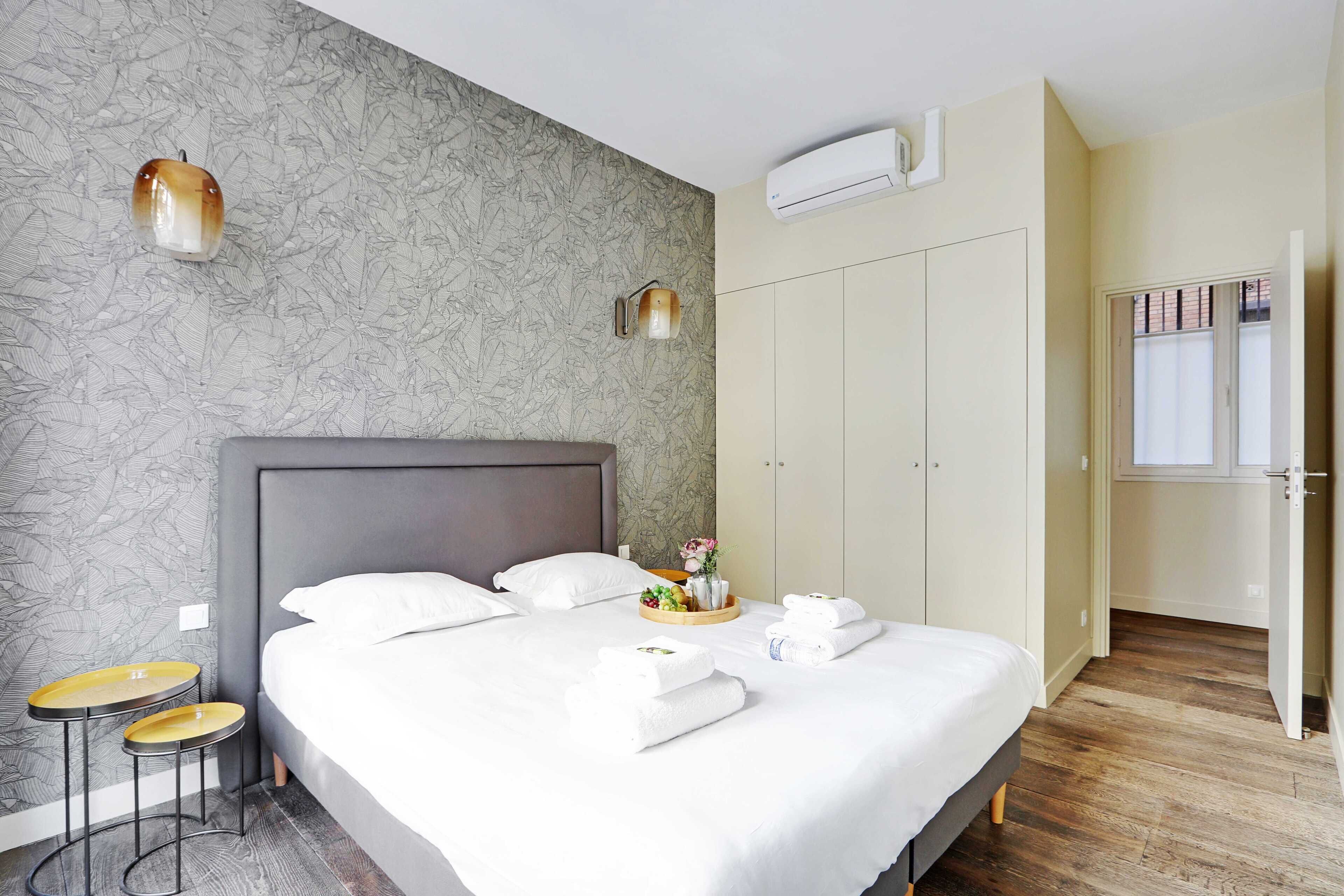 A modern hotel room with a large bed, two side tables, a wardrobe, and an air conditioning unit.