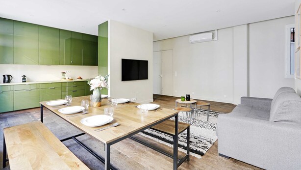 A modern kitchen with green cabinets, a dining table set for four, and a grey sofa in the living area. - Appartement Beaubourg (Paris)