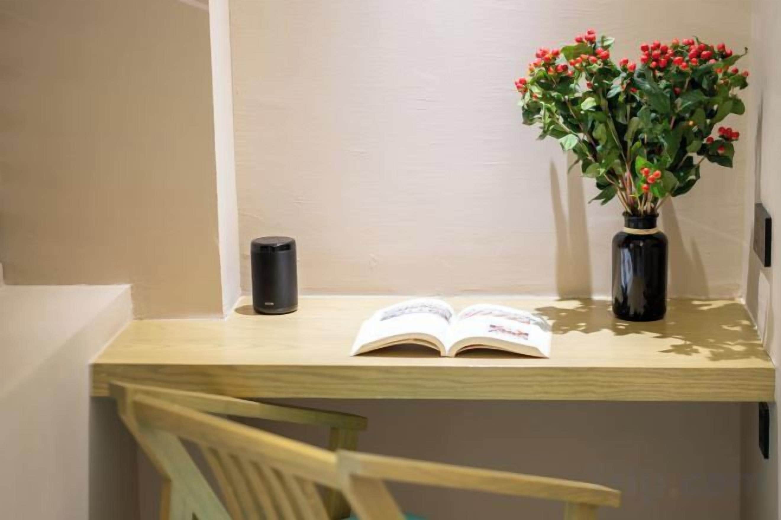 Un escritorio de madera con un libro, un altavoz negro y una jarrón con flores rojas.