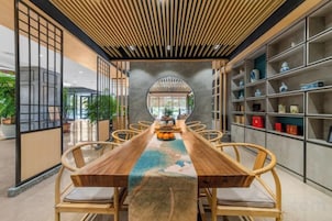 Uma longa mesa de jantar de madeira com cadeiras, rodeada por prateleiras com objetos decorativos.
