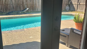 Uitzicht door een glazen deur op een zwembadgedeelte met een houten omheining, tuinmeubels en een helder blauw zwembad.
