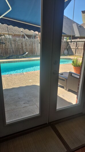 A view through a glass door of a pool area with a wooden fence, outdoor furniture, and a clear blue pool.