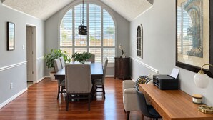 A spacious room with a high ceiling, a dining area with a wooden table and chairs, a large window with blinds, and a framed artwork on the wall. - House of blue! Beautiful pool, near IAH airport (Humble)