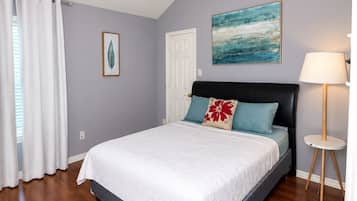 A bedroom with a bed, a ceiling fan, a door, a window with curtains, a painting, and a lamp.