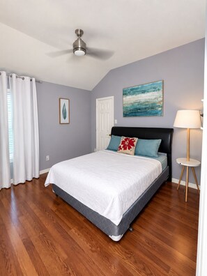 Un dormitorio con cama, ventilador de techo, puerta, ventana con cortinas, un cuadro y una lámpara.