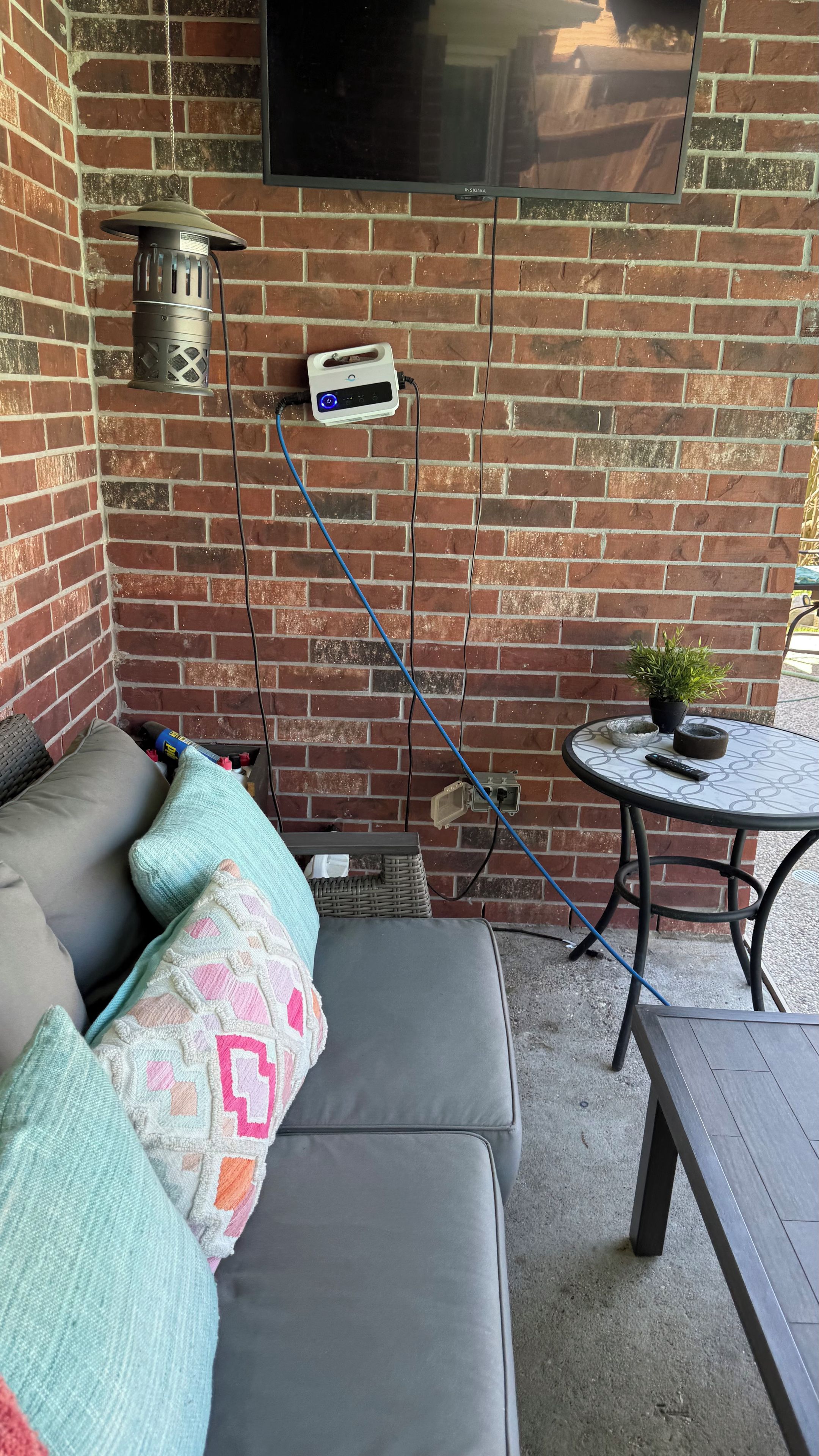 Eine Terrasse mit einer Ziegelwand, einem grauen Sofa mit gemusterten Kissen, einem kleinen runden Tisch und einem Topfpflanzen.
