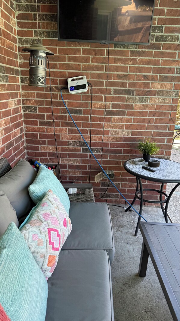 A patio area with a brick wall, a gray sofa with patterned cushions, a small round table, and a potted plant.