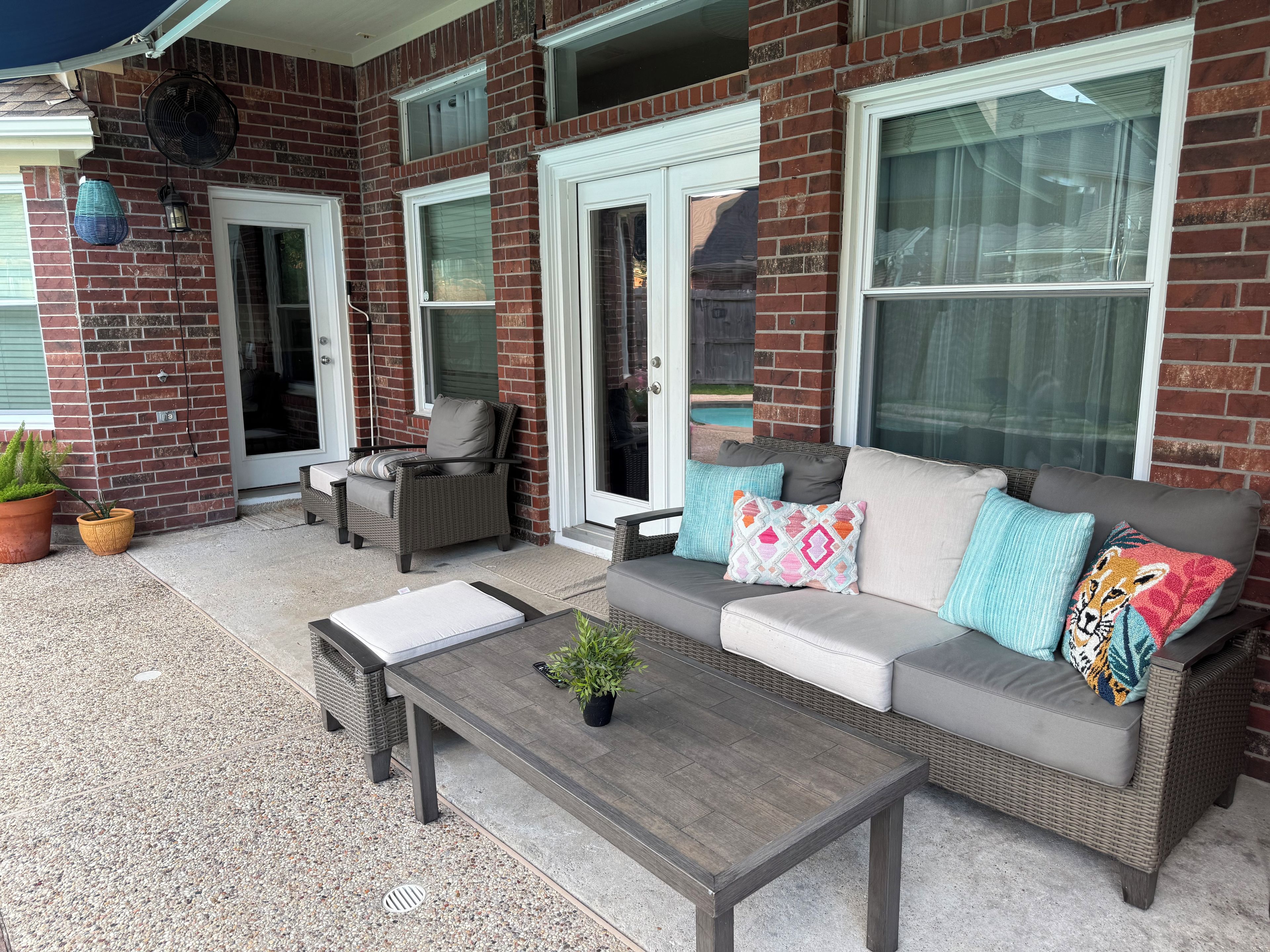 A patio area with a grey sofa, a wicker chair, and a small table. There are potted plants and a fan mounted on the wall.