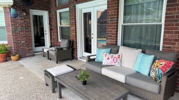 A patio area with a grey sofa, a wicker chair, and a small table. There are potted plants and a fan mounted on the wall.