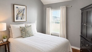 A bedroom with a bed, a nightstand, a lamp, a framed picture, a window with curtains, and a dark wooden cabinet. - House of blue! Beautiful pool, near IAH airport (Humble)