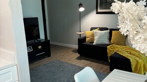A modern living room with a flat-screen TV, a black sofa with yellow cushions, a white vase with white flowers, and a grey rug.