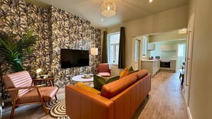 Ein Wohnzimmer mit Sofa, Sessel und Fernseher, einer Wand mit Muster und einer Küche im Hintergrund.