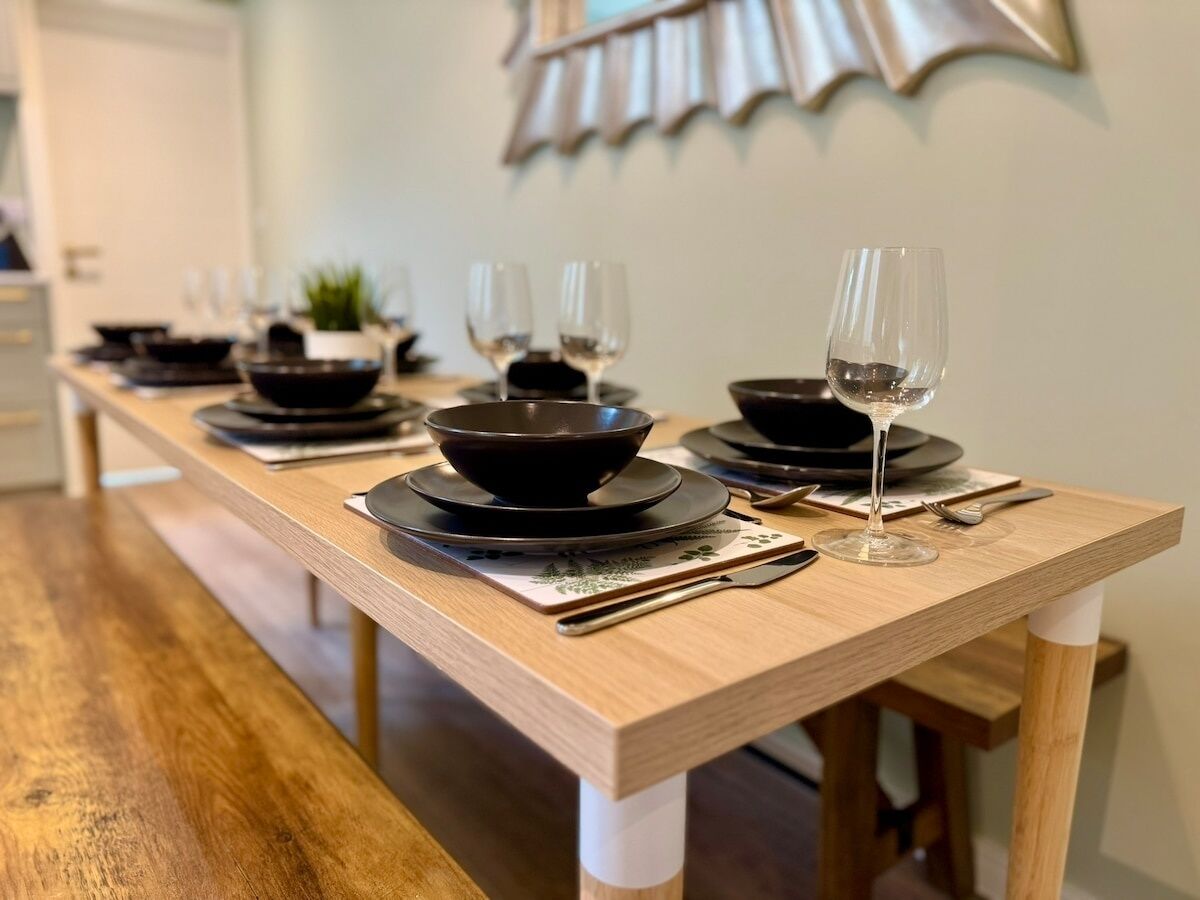 Une table à manger dressée avec des bols, des assiettes et des verres à vin noirs, et un banc en bois en premier plan.