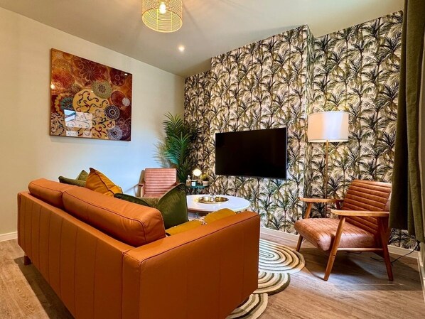 Una sala de estar con un sofá, una silla, un televisor de pantalla plana, una lámpara y un papel tapiz estampado.